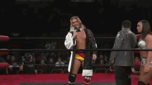 a man stands in a wrestling ring talking into a microphone while two women stand behind him