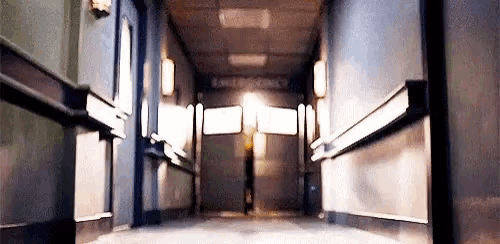 a person is walking down a hallway in a hospital with a light shining through the doors .