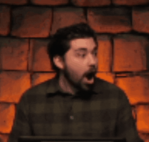 a man with a beard is sitting in front of a brick wall covering his mouth with his hand .