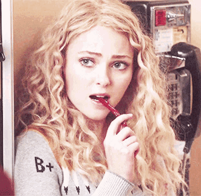 a woman with curly blonde hair is holding a red object in her mouth and wearing a sweater with the letter b on it