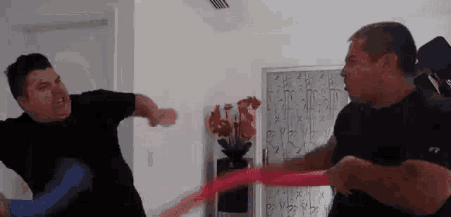 two men are fighting each other in a living room while holding a red ribbon .