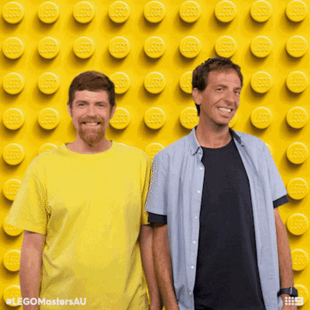 two men are standing in front of a wall of yellow lego bricks
