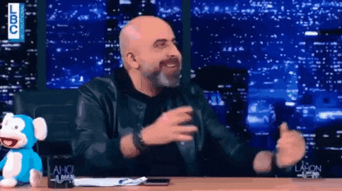 a bald man with a beard is sitting at a table with a stuffed animal and giving a thumbs up .
