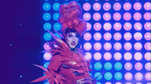 a drag queen is standing in front of a wall of lights and balloons .
