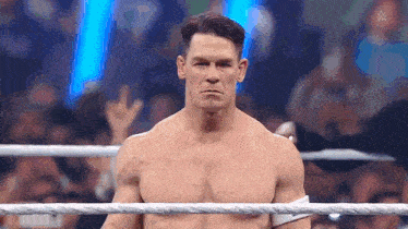 a shirtless wrestler is standing in a wrestling ring with a crowd behind him .