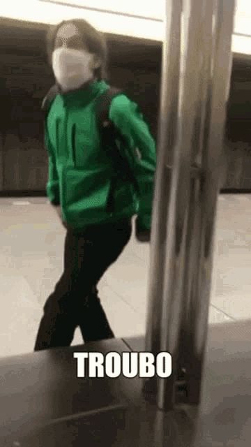 a man wearing a mask and a green jacket is standing next to a pole with the words troubo written on it .