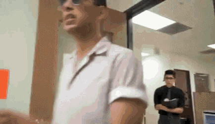 a man wearing sunglasses is standing in a hallway with another man standing behind him holding a clipboard .