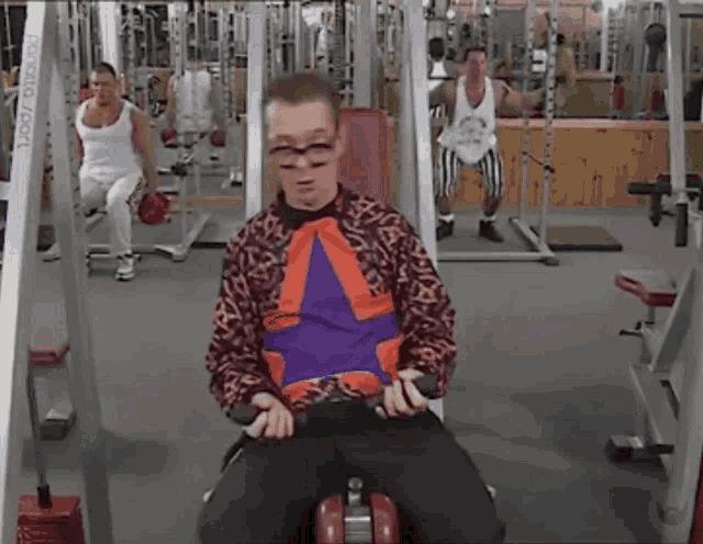 a man in an orange and purple shirt is sitting on a machine in a gym with a sign that says ' reebok '