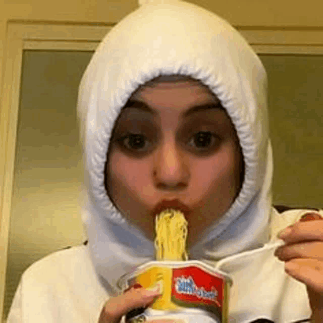 a woman wearing a white hooded hat is eating noodles from a cup .