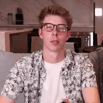a young man wearing glasses and a floral shirt is sitting on a couch in a living room .