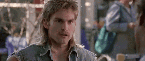 a man with long hair and a mullet is wearing a denim vest and looking at the camera .
