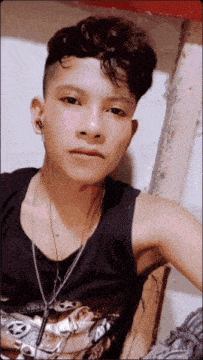 a young man wearing a black tank top and a necklace with the letter a on it looks at the camera