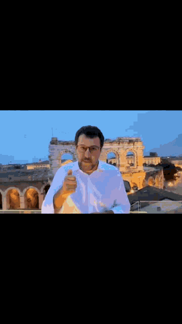 a man wearing glasses and a white shirt is giving a thumbs up in front of a ruined building .