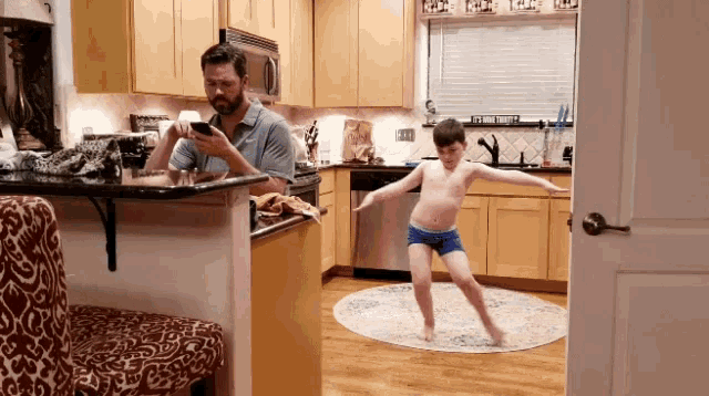 a man is looking at his phone while a boy is dancing in a kitchen