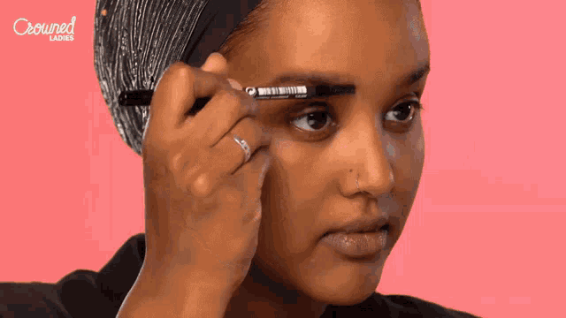 a woman is applying mascara to her eyebrows and the words crowned ladies are visible in the background