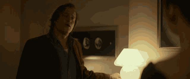 a man in a brown jacket stands next to a lamp in a dark room