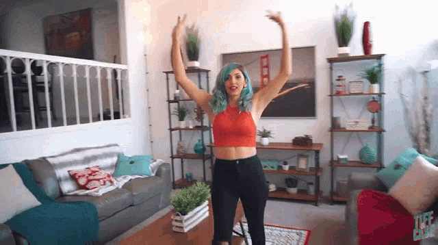 a woman with blue hair is dancing in a living room with her arms in the air .
