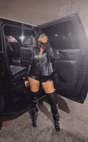 a woman in leather shorts and thigh high boots stands in front of a car