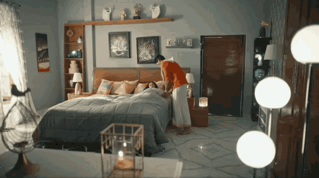 a woman standing next to a bed in a bedroom with lamps on the floor