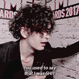 a man with curly hair says " you used to say that i was shit " in front of a brick wall
