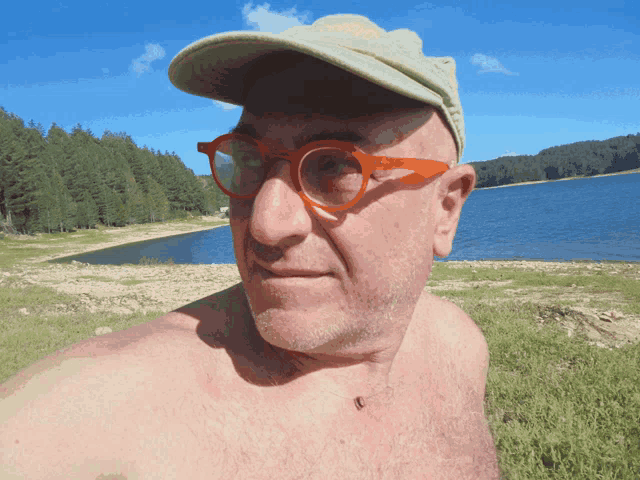 a shirtless man wearing glasses and a hat stands near a lake