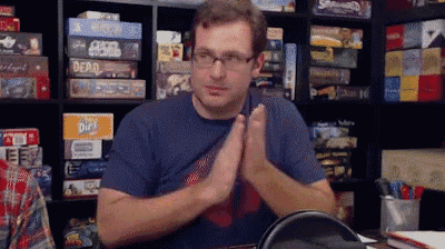 a man wearing glasses is sitting at a table with his hands folded in front of a shelf full of board games .