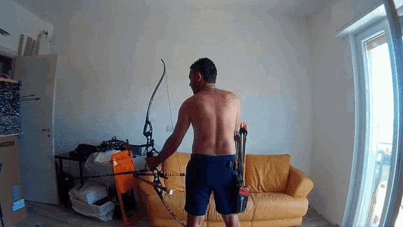 a man without a shirt is holding a bow and arrow in front of a yellow couch