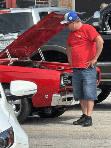 a man in a red shirt is looking at a red car with the hood open