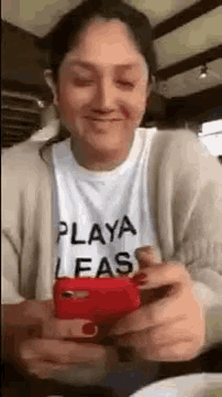 a woman wearing a t-shirt that says `` playa leas '' is holding a cell phone in her hands .