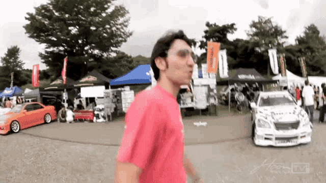 a man in a red shirt is walking in a parking lot with a car parked in the background