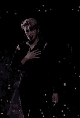 a man in a black jacket is standing on a stage holding his hand to his forehead .