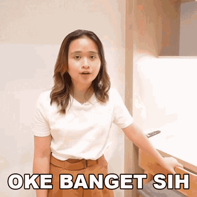 a woman wearing a white shirt and brown pants is standing in front of a desk and says oke banget sih