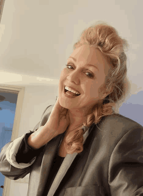 a woman wearing a grey suit jacket is smiling and touching her neck