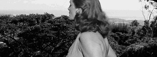 a black and white photo of a woman standing in front of a lush green forest .