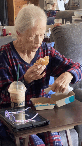an elderly woman is sitting at a table eating a sandwich and drinking a coffee with the letter ea on it