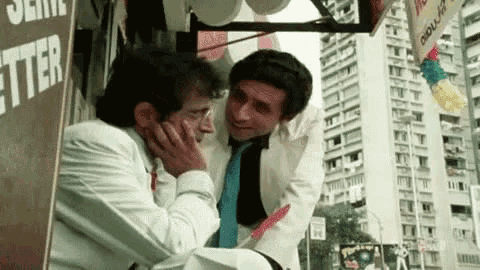 two men are standing next to each other in front of a building and talking .