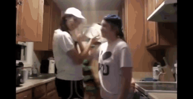 two girls are standing in a kitchen with one wearing a shirt that says ' u ' on it .