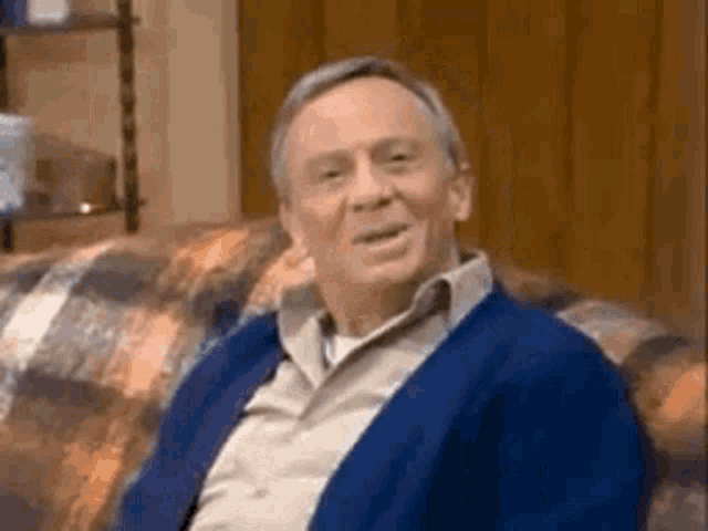 an elderly man is sitting on a couch wearing a blue cardigan and smiling .