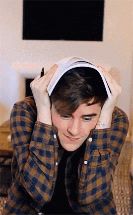 a young man in a plaid shirt holds a book to his head