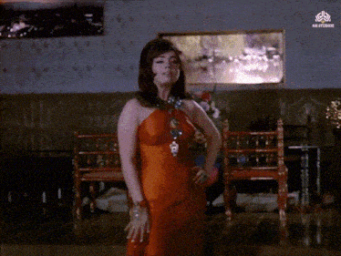a woman in an orange dress is standing in a room with a nb studios logo on the corner