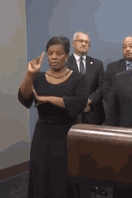 a woman in a black dress is giving a speech in front of a group of men in suits