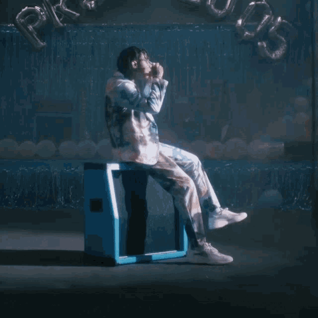 a man in a suit sits on a blue box with balloons in the background
