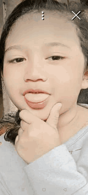 a little girl is sticking her tongue out while holding her hand to her chin .