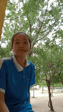 a girl in a blue shirt with the letter a on it stands in front of a tree