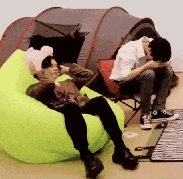two men are sitting on a bean bag chair in front of a tent