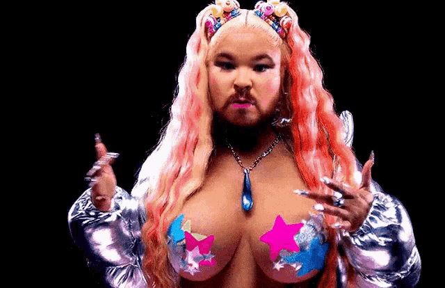 a woman with a beard and pink hair is wearing a silver jacket and pink stars on her breasts .