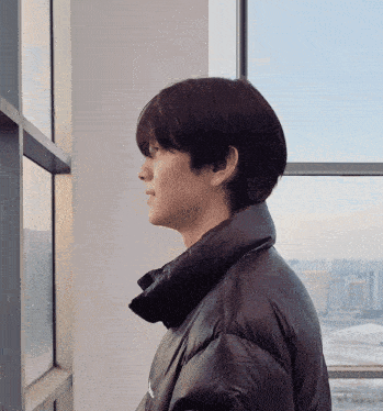a man in a black jacket stands in front of a window looking out