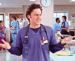 a man wearing a blue scrub top with a stethoscope around his neck stands in a hospital room