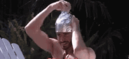 a shirtless man is washing his hair with a tin foil hat while sitting in a chair .