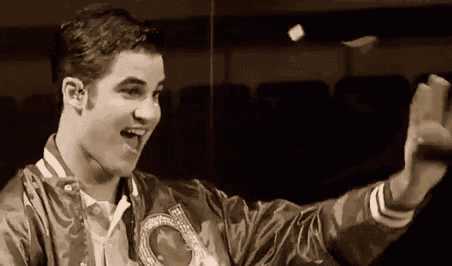 a young man in a varsity jacket is making a funny face and waving his hand in a black and white photo .